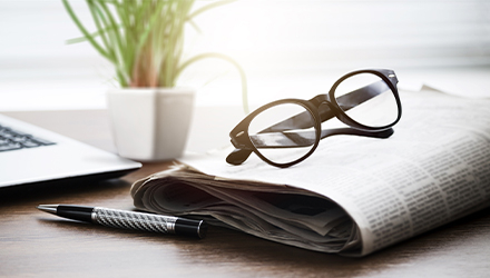 Newspaper, glasses, pen, and laptop on desk